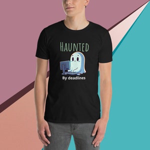 以下が含まれることがあります： 黒いTシャツに、コンピューターに座っている白い幽霊と「HAUNTED By deadlines」というテキストが書かれています。