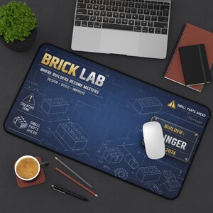 以下が含まれることがあります： 「BRICK LAB」の文字と、設計図風のイラストが描かれた、青と黒の長方形のマウスパッド。マウスパッドには「WHERE BUILDERS BECOME MASTERS」と「DESIGN - BUILD - IMPROVE」の文字も。白いマウスが上に置かれています。