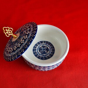 May include: A blue and white ceramic trinket box with a lid. The lid has a gold-colored knob. The box is decorated with intricate floral patterns.