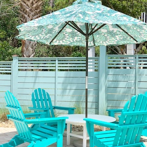 May include: Four turquoise adirondack chairs surrounding a white round table with a patterned umbrella overhead. The chairs and table are on a brick patio.
