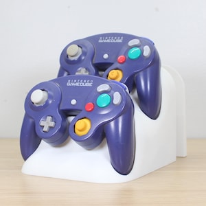 Puede incluir: Dos mandos morados de Nintendo GameCube con botones coloridos se muestran en un soporte blanco. Los mandos tienen un diseño clásico con una cruceta, una palanca analógica y varios botones. El soporte es blanco y tiene un diseño sencillo.