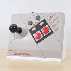 May include: A grey Nintendo NES Advantage arcade-style joystick controller featuring a black ball-top stick, two large red action buttons, two smaller grey turbo dials, and white buttons labeled Select and Start, with the red Nintendo NES Advantage logo.