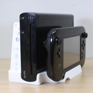 May include: A black Wii console, a black Wii U GamePad, and a white Wii remote are displayed in a white stand. The Wii U GamePad has a screen and buttons. The Wii remote has buttons and the Wii logo.