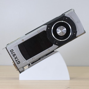 May include: A silver and black GTX 970 graphics card, angled on a white stand. The card has a large fan and the text "GTX 970" is visible. The stand is a simple, modern design, and the background is a neutral color.