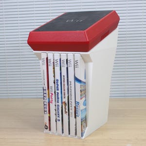 Peut inclure: Un support de rangement en plastique blanc contenant six boîtiers de jeux vidéo Wii. Le support présente une section supérieure rouge avec un panneau noir affichant le logo Wii. Les boîtiers sont disposés verticalement, montrant des titres comme Wii Party, Super Mario Galaxy 2, Super Smash Bros. Brawl, Mario Kart Wii et Wii Sports Resort.