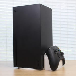 Könnte beinhalten: Eine mattschwarze, rechteckige Videospielkonsole, die vertikal neben einem schwarzen kabellosen Gamecontroller steht. Der Controller ruht auf einem weißen Kunststoffständer. Die Konsole verfügt über einen runden Einschaltknopf und einen Schlitz.