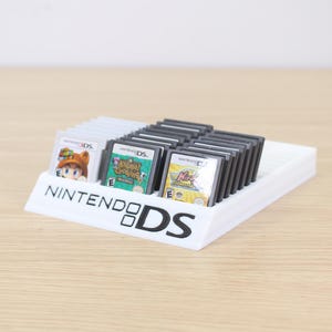Può includere: Un supporto bianco per cartucce di giochi Nintendo DS con diverse cartucce di gioco. Il supporto presenta il logo Nintendo DS. Sono visibili diverse cartucce di gioco, tra cui Animal Crossing e altri titoli.