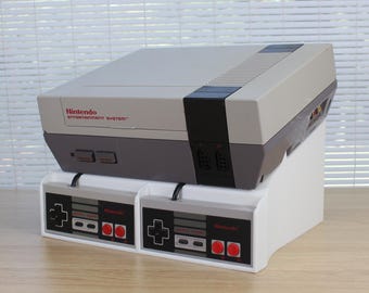 May include: A gray and black Nintendo Entertainment System console resting on a white plastic stand. Two rectangular gray controllers with black directional pads and red buttons are stored in the front slots of the stand.