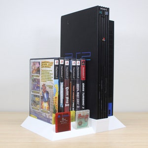 Könnte beinhalten: Eine schwarze PlayStation 2 Konsole mit einem weißen Ständer, der einen Stapel PlayStation 2 Spielhüllen hält. Der Ständer hält auch ein paar Speicherkarten und eine PlayStation 2 Spielhülle.