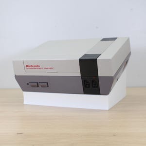 May include: A classic Nintendo Entertainment System (NES) console in gray and black, with the Nintendo logo in red. The console is on a white stand, showcasing its iconic design. A retro gaming item.