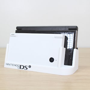 Puede incluir: Un soporte de carga blanco para Nintendo DS con una Nintendo DS negra en su interior. El soporte tiene el logotipo de Nintendo DS en la parte delantera.