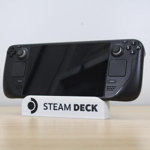 May include: A black Steam Deck handheld gaming console resting on a white stand. The stand has the words "STEAM DECK" printed on it in black. The console features a large screen and multiple buttons.