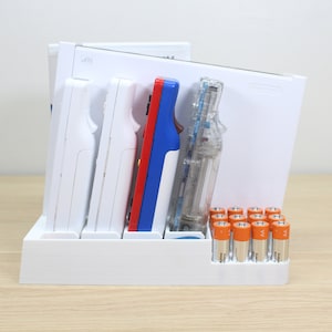 Puede incluir: Un soporte de pl&aacute;stico blanco con ranuras para guardar mandos de videojuegos, una caja de pl&aacute;stico transparente y una pila de pilas AA. El soporte tiene una etiqueta que dice "Wii".