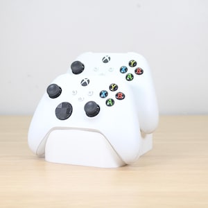 Peut inclure: Deux manettes Xbox blanches sur un socle de chargement blanc. Les manettes ont des joysticks noirs et des boutons colorés A, B, X et Y. Le socle est posé sur une surface en bois clair.