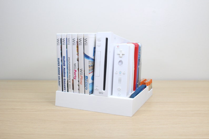 Puede incluir: Soporte de pl&aacute;stico blanco con una consola Wii blanca, un mando Wii blanco y una pila de discos de juego de Wii. Los discos incluyen t&iacute;tulos como "Wii Play", "Wii Party", "Mario Kart Wii" y "Super Mario Galaxy".