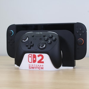 May include: A black Nintendo Switch console with a black controller and a black controller stand. The stand has the Nintendo Switch logo in red and white. The console is on a light brown surface.