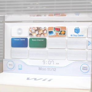 Op de afbeelding: Een witte Wii-console-interface met verschillende kanaalpictogrammen, waaronder Weer, Nieuws en Wii Shop. Het scherm toont de tijd als 9:07 uur en de datum als ma 11/12. De console is ingesloten in een doorzichtige plastic hoes.
