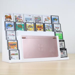 May include: White plastic game organizer holding a rose gold Nintendo DS and multiple game cartridges. The cartridges are in black cases with colorful game labels. The organizer is on a light brown surface.