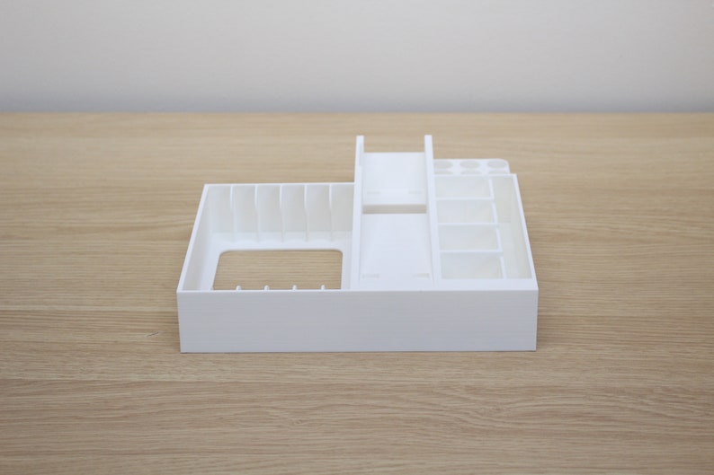 Puede incluir: Organizador de pl&aacute;stico blanco con m&uacute;ltiples compartimentos para guardar objetos peque&ntilde;os. El organizador tiene un compartimento rectangular grande en la parte delantera y un compartimento m&aacute;s peque&ntilde;o con divisores verticales en la parte trasera.
