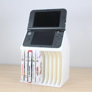 Può includere: Supporto in plastica bianca con fessure per riporre le cartucce di gioco Nintendo 3DS. Una console di gioco portatile Nintendo 3DS nera è posizionata sopra il supporto.