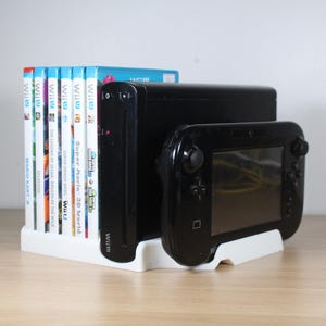 Può includere: Una console Wii U nera con un gamepad e diverse custodie di giochi. La console e il gamepad sono neri e le custodie dei giochi hanno etichette colorate. Gli articoli sono disposti in un supporto bianco su una superficie di legno.