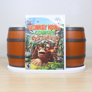 Puede incluir: Un soporte de consola de juegos Nintendo Wii blanco con dos barriles marrones a cada lado. El soporte sostiene una caja de juego de video para Donkey Kong Country Returns para Wii.
