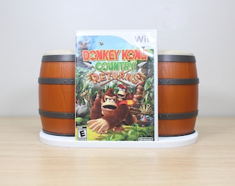 May include: A white Nintendo Wii game console stand with two brown barrels on either side. The stand is holding a video game case for Donkey Kong Country Returns for the Wii.