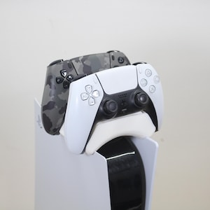 Op de afbeelding: Een witte en zwarte PlayStation 5 console standaard met twee gamecontrollers. Eén controller is wit met zwarte accenten, en de andere is camouflage. De standaard is wit met een zwarte basis.