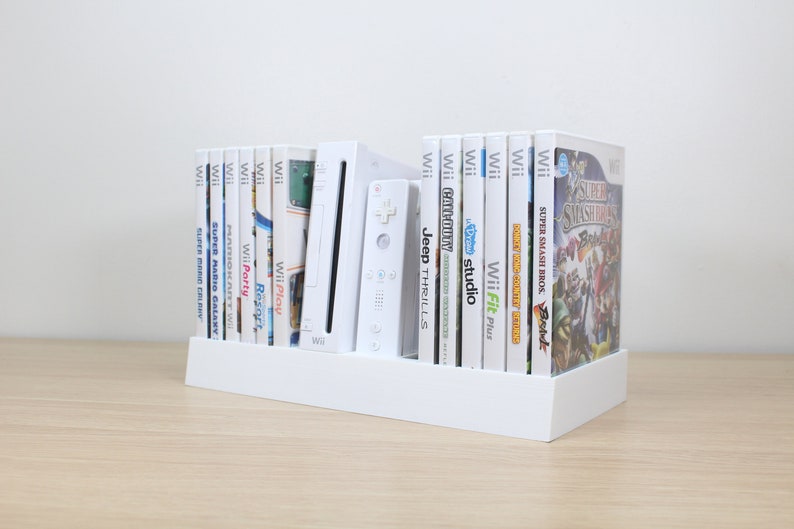 Puede incluir: Una consola Wii blanca con un soporte blanco que sostiene varios cartuchos de juegos de Wii. Los cartuchos est&aacute;n organizados verticalmente e incluyen t&iacute;tulos como "Wii Play", "Wii Party", "Super Mario Galaxy", "Super Smash Bros. Brawl" y "Wii Fit Plus".