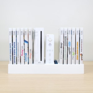 Puede incluir: Una consola Wii blanca y un mando a distancia Wii blanco se muestran en un estante blanco con una colecci&oacute;n de estuches de juegos Wii blancos. Los t&iacute;tulos de los juegos incluyen Super Mario Galaxy, Mario Kart Wii, Wii Party, Wii Play, Wii Resort, Jeep Thrills, Wii Fit Plus y Super Smash Bros. Brawl.