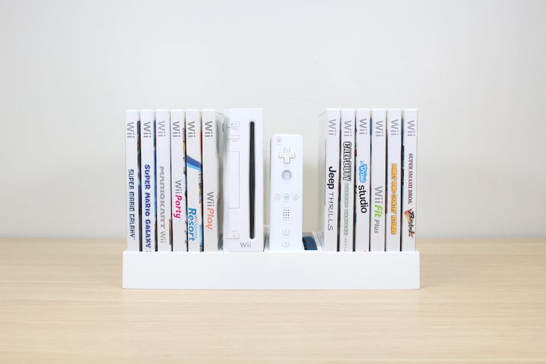 Puede incluir: Una consola Wii blanca y un mando a distancia Wii blanco se muestran en un estante blanco con una colecci&oacute;n de estuches de juegos Wii blancos. Los t&iacute;tulos de los juegos incluyen Super Mario Galaxy, Mario Kart Wii, Wii Party, Wii Play, Wii Resort, Jeep Thrills, Wii Fit Plus y Super Smash Bros. Brawl.