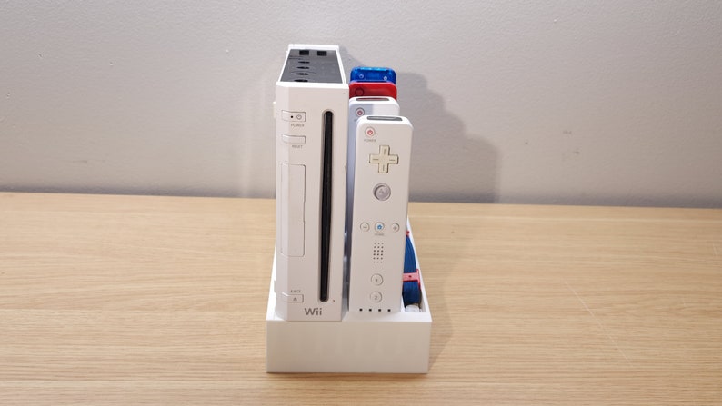 Puede incluir: Una consola Nintendo Wii blanca con un soporte blanco y cuatro mandos Wii blancos. La consola tiene un panel frontal negro con la palabra "Wii" impresa en &eacute;l. Los mandos tienen botones y correas de varios colores.