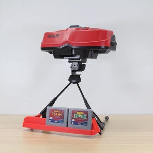 以下が含まれることがあります： 赤と黒のバーチャルボーイビデオゲーム機と2つのゲームカートリッジ。コンソールは、赤いベースの黒い三脚スタンドに取り付けられています。「VIRTUAL BOY」の文字がコンソールに表示されています。