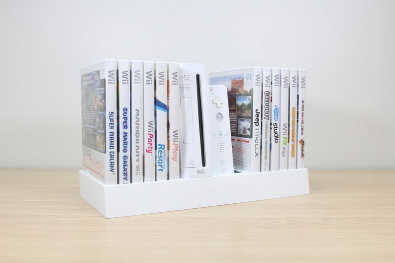 Puede incluir: Una consola Wii blanca con un soporte blanco que contiene una colecci&oacute;n de discos de juegos de Wii. Los t&iacute;tulos de los juegos incluyen "Super Mario Galaxy", "Mario Kart Wii", "Wii Party", "Wii Play", "Wii Resort", "Wii Fit Plus", "Studio", "Cadouy", "Jeep Thrills" y "Super Smash Bros. Brawl".