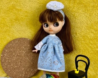 Puede incluir: Una muñeca con un vestido de encaje azul claro con ribetes de encaje blanco y una diadema blanca con una perla. La muñeca está de pie sobre un posavasos de corcho con una figurita de pez de colores adornada con joyas. Una pequeña silla negra con un pato de goma amarillo en el asiento está en el fondo.