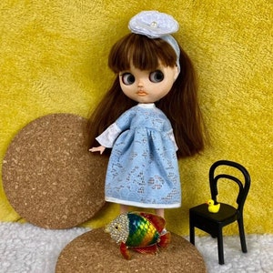 Può includere: Una bambola che indossa un abito di pizzo blu chiaro con bordi in pizzo bianco e una fascia bianca con una perla. La bambola è in piedi su un sottobicchiere di sughero con una statuetta di pesce colorata e decorata con gioielli. Una piccola sedia nera con un'anatra di gomma gialla sul sedile è sullo sfondo.