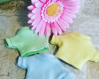 Puede incluir: Tres camisas de punto en miniatura en colores pastel: verde claro, amarillo y azul. Las camisas están dispuestas sobre una superficie texturizada con una flor rosa en el fondo.