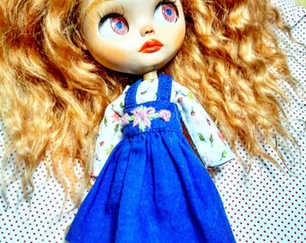 May include: A doll with long, curly, red hair wearing a blue denim overall dress with a floral embroidery design. The doll has blue eyes with pink accents and is wearing a white shirt with a floral print.