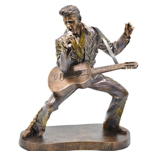 May include: A bronze-effect statue of a male figure playing a guitar and singing into a microphone. The figure is wearing a detailed jumpsuit with gold accents and a guitar strap. The statue is on a rectangular base.