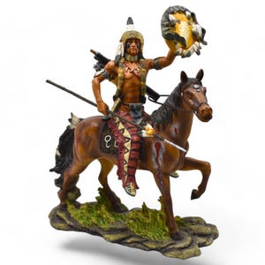 May include: A figurine of a Native American warrior riding a brown horse, holding a feathered drum and a spear. The figurine is wearing a feathered headdress and a colourful loincloth. The horse has a saddle and a blanket.