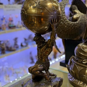 May include: A bronze Atlas statue kneeling and supporting a globe. The globe is detailed with zodiac symbols and celestial designs. The statue is on a rocky base, and the overall aesthetic is classical and detailed.