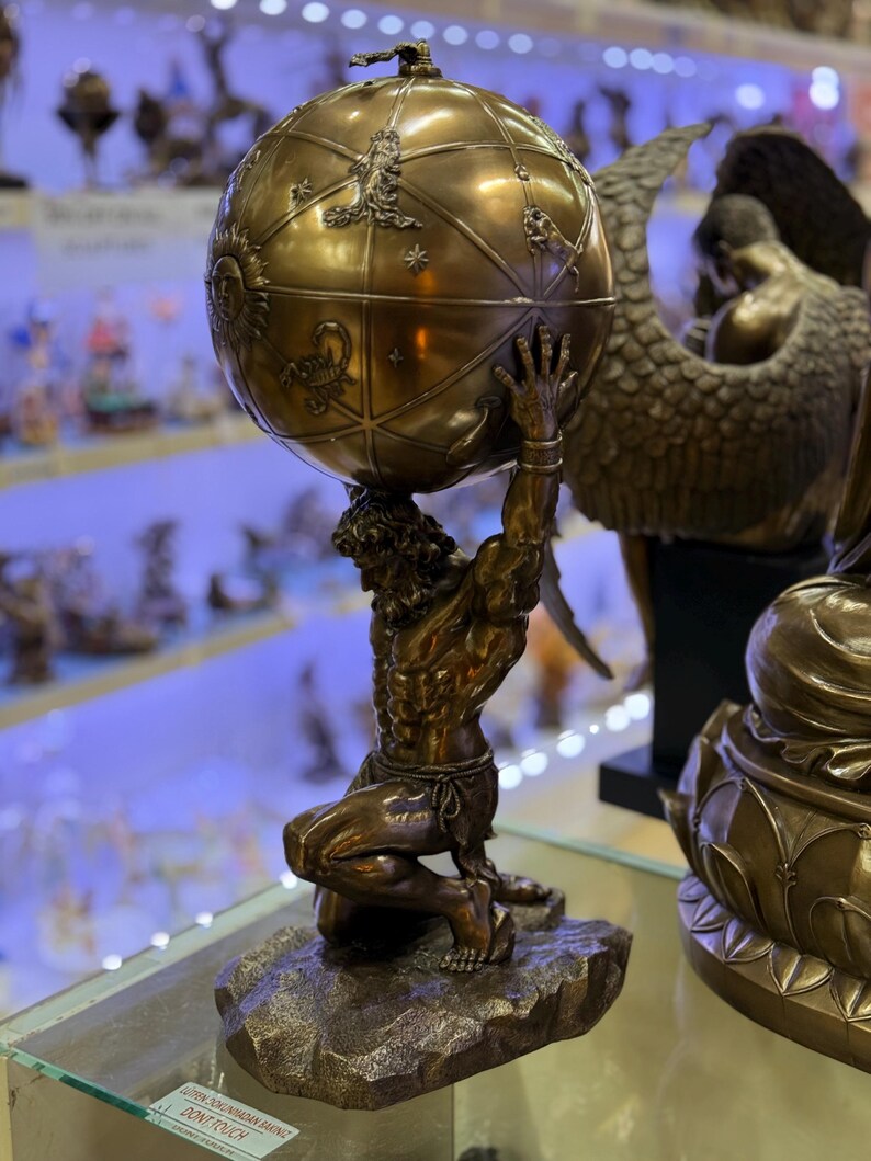 May include: A bronze Atlas statue kneeling and supporting a globe. The globe is detailed with zodiac symbols and celestial designs. The statue is on a rocky base, and the overall aesthetic is classical and detailed.