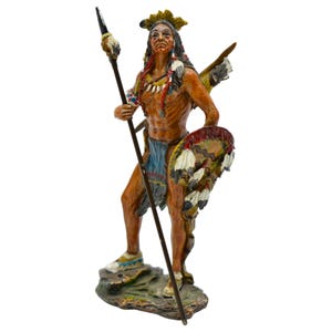 May include: A hand-painted figurine of a Native American warrior holding a spear and shield. The warrior is wearing a feathered headdress and a colourful loincloth. The figurine is standing on a base with a rocky texture.