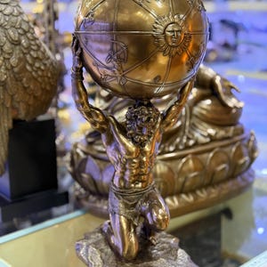 May include: A bronze sculpture of Atlas kneeling and supporting a globe. The globe features detailed depictions of constellations and a sun face. The sculpture is on a rocky base and has a metallic finish.