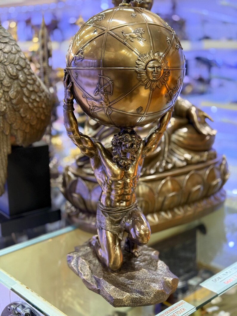 May include: A bronze sculpture of Atlas kneeling and supporting a globe. The globe features detailed depictions of constellations and a sun face. The sculpture is on a rocky base and has a metallic finish.