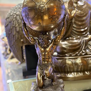 May include: A bronze statue of Atlas kneeling and supporting a globe. The globe features zodiac symbols and a sun face. The statue is on a stone base, with a Buddha statue in the background.