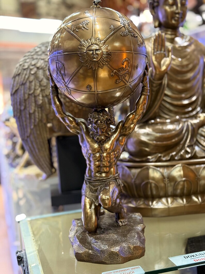 May include: A bronze statue of Atlas kneeling and supporting a globe. The globe features zodiac symbols and a sun face. The statue is on a stone base, with a Buddha statue in the background.