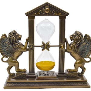 May include: A gold-toned hourglass with winged lion statues on either side, set on a decorative base. The hourglass is made of glass and has a clear top and a yellow bottom.