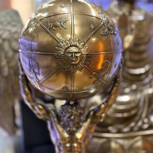 May include: A bronze sculpture of Atlas holding a celestial globe. The globe features zodiac symbols and a sun face. The sculpture is detailed with a metallic finish, showcasing the figure's muscular form and the globe's intricate design.