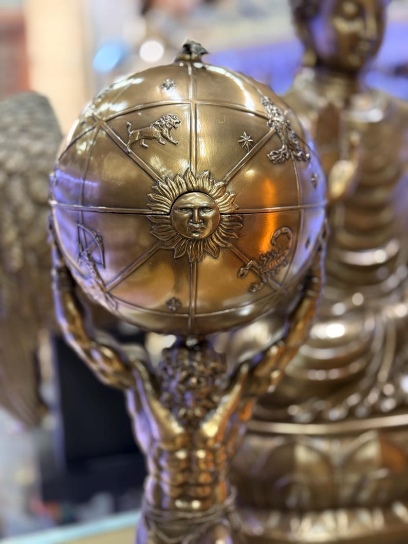 May include: A bronze sculpture of Atlas holding a celestial globe. The globe features zodiac symbols and a sun face. The sculpture is detailed with a metallic finish, showcasing the figure's muscular form and the globe's intricate design.